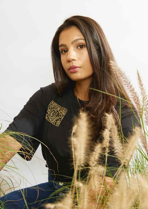 Woman sitting amongst grasses wearing the rose embroidery in a square on the upper right of the black Asmuss Evelyne Tshirt that has been hand embellished by Asmuss designer