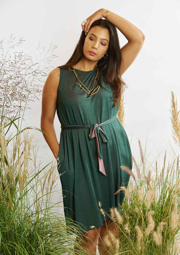 Woman with a hand over her head and the other in the pocket of the Asmuss Drift Dress that is belted with the self fabric belt while standing amongst grasses