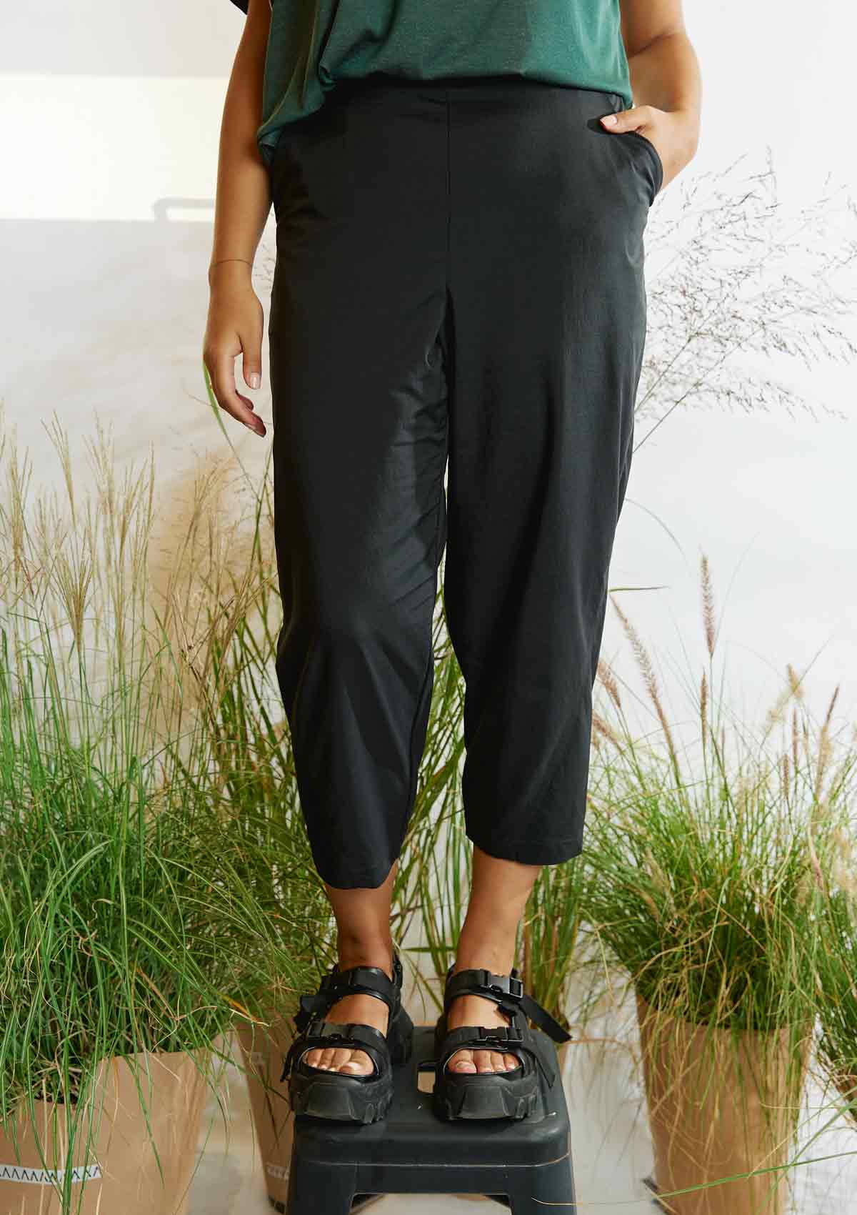 Woman standing on a stool amongst grasses with a hand in a pocket of the Asmuss Oval Trousers and wearing balck sandals