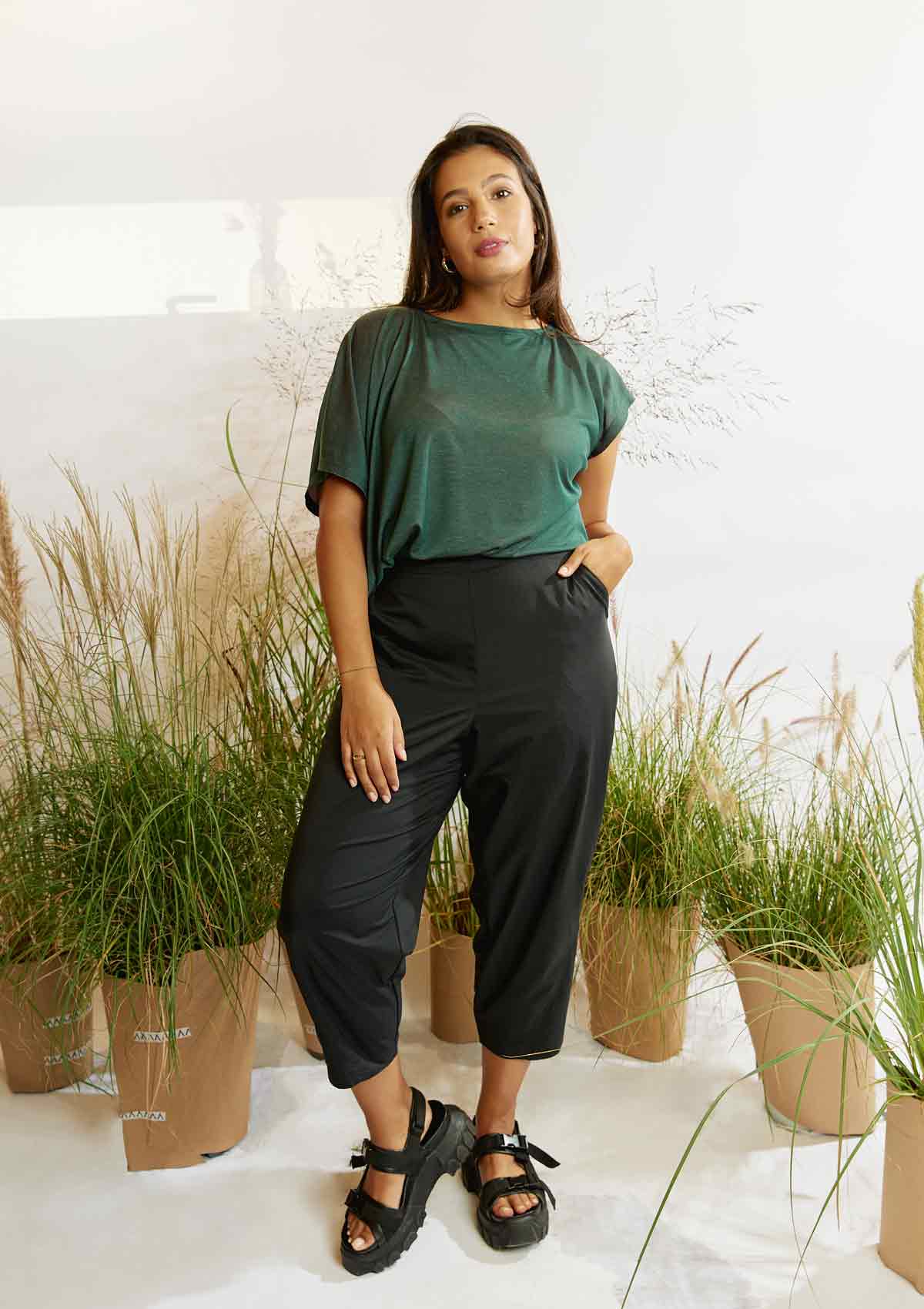 Woman standing amongst grasses with one hand in a pocket wearing the pine green Heed T-shirt and the black Asmuss Oval Trousers