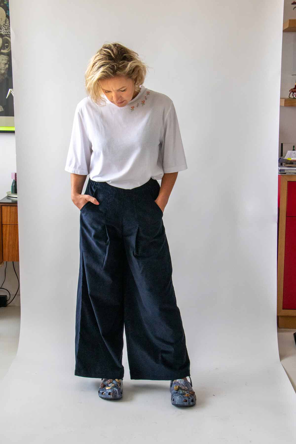 Woman, with short wavy blond hair, looking down and wearing the Asmuss Ridge Trousers in Navy and Asmuss Evelyn T-shirt in White, with her hands in the trousers pockets