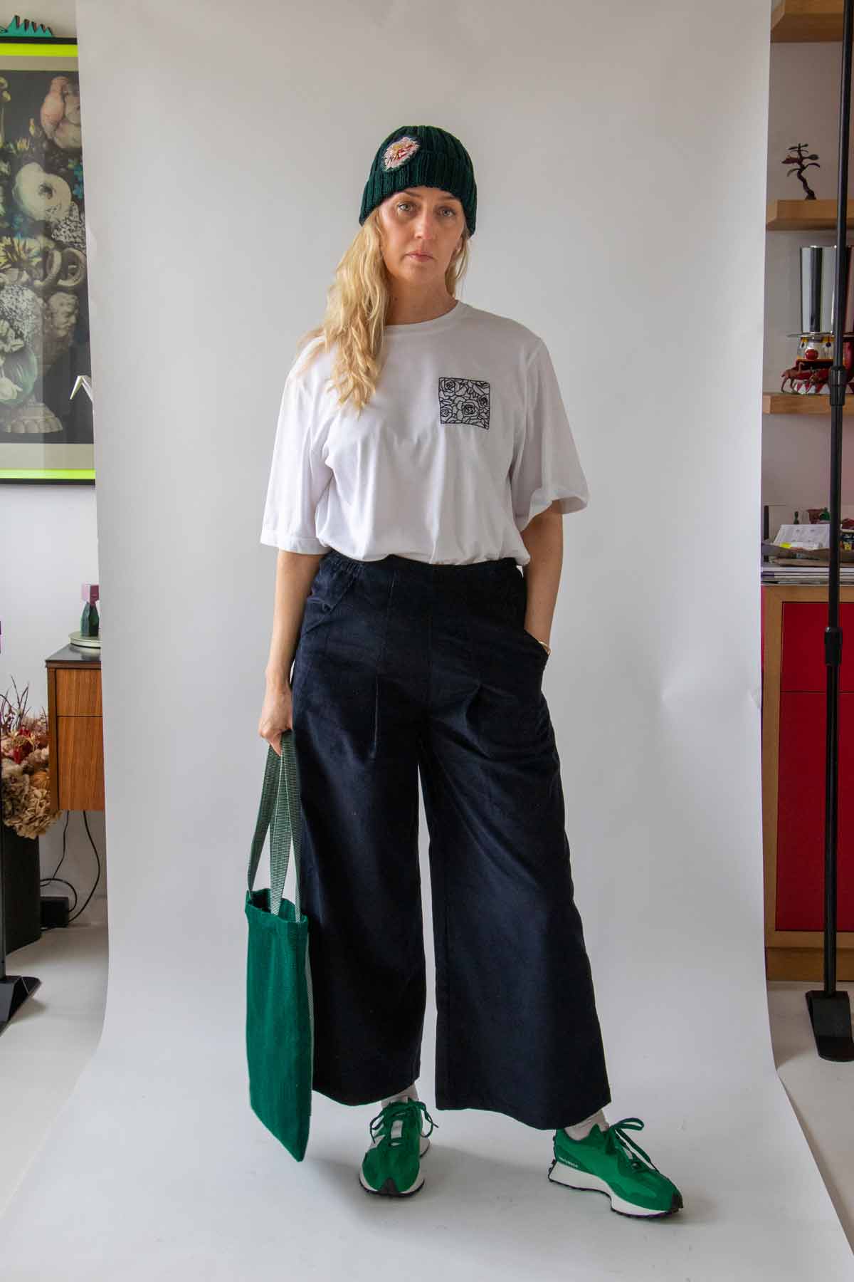 Woman, with long wavy blond hair, standing front on wearing the Asmuss Ridge Cords in Navy, Evelyn T-shirt in White and Hand knitted Beanie holding a green bag in her right hand