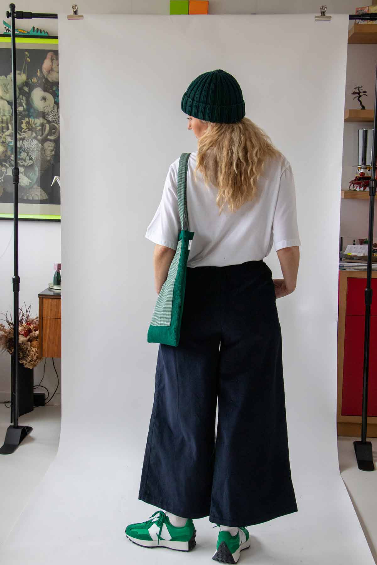 Woman, with long wavy blond hair, standing back on wearing the Asmuss Ridge Cords in Navy, Evelyn T-shirt in White and Hand knitted Beanie with a green bag on her right shoulder