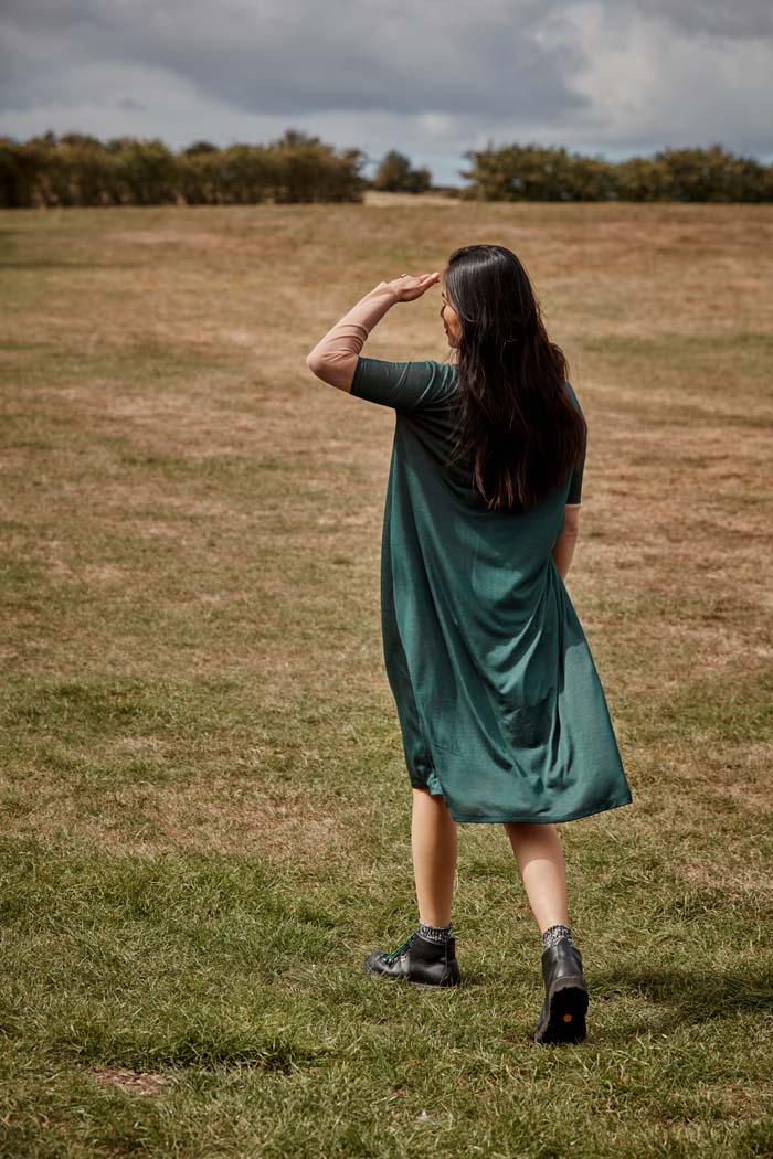 Woman walking away wearing the fluid moving Asmuss Asymmetric Pleat Dress in Pine Green. Slim arms, loose fitting body with pleat detail and pocket it makes a great dress for your everyday adventures. Made from a blend of recycled polyester and Tencel a great sustainable fabric. A stylish, sustainable addition to the modern woman's wardrobe.