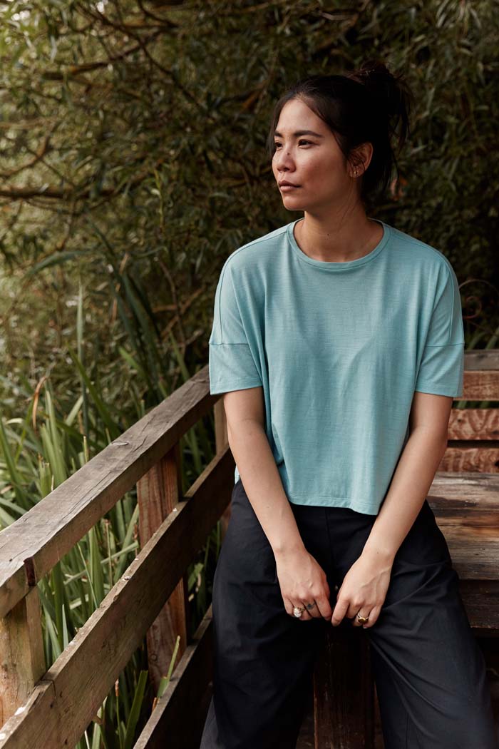 Woman sitting on a wooden bench in the countryside wearing the Asmuss Boxy Cropped T-shirt in Sea Green. Made in the UK from a blend of wool and 37.5 Technology to help keep you at the ideal body temperature. One of the best elevated essentials for the modern woman's everyday adventures.