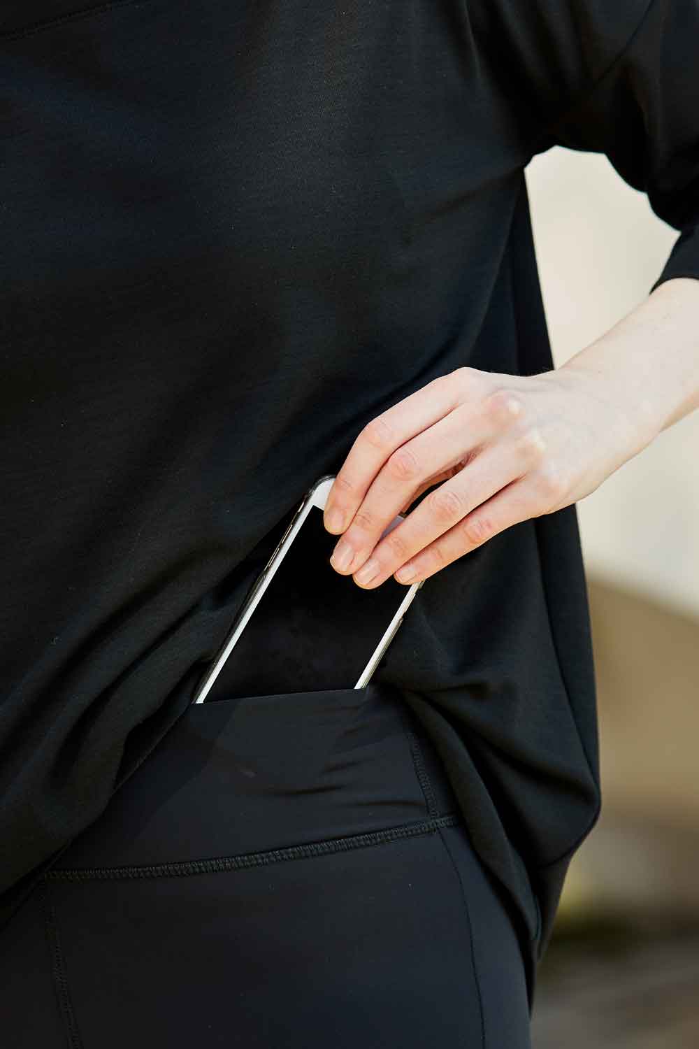 Showing how the kangaroo pocket in the waistband of the Asmuss Twisted Leggings holds a phone. The high waisted Asmuss Twisted Leggings with zip detail on lower front leg, pocket in front waistband and drawstring to keep them snug. Made in the UK from recycled nylon and elastane for 4 way stretch. Sustainable activewear that looks great on for all your everyday adventures including lazing on the couch or out running.
