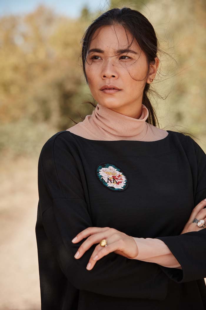 Woman wearing the Asmuss Oversized Boat Neck Sweat in black with embroidered rose badge. Stylish, sustainable lifestyle womenswear for any of your everyday adventures whether they are everyday or when you are travelling