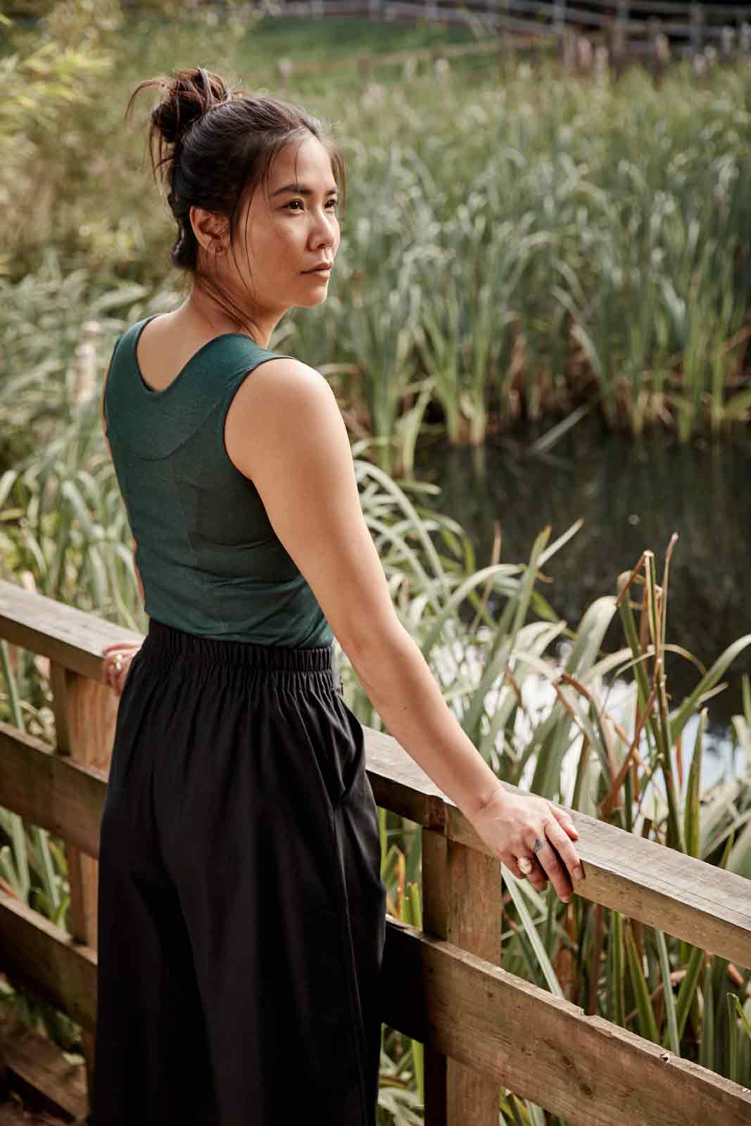 Back view of woman wearing the Asmuss Panelled Tank in Pine Green leaning on a railing near a pond filled with reeds. Stylish and sustainable lifestyle womenswear for your everyday adventures and activewear.