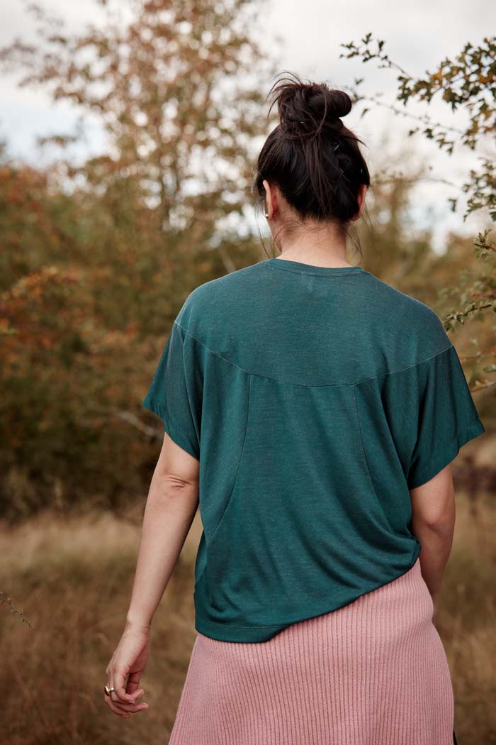 Back view of woman in the outdoors wearing the Asmuss Panelled T-shirt in Pine Green. Made from a sustainable fabric that feels luxurious and drapes. A t-shirt elevated with interesting detailing. A great stylish and sustainable luxury t-shirt that is the perfect activewear t-shirt that can be worn on your everyday adventures.
