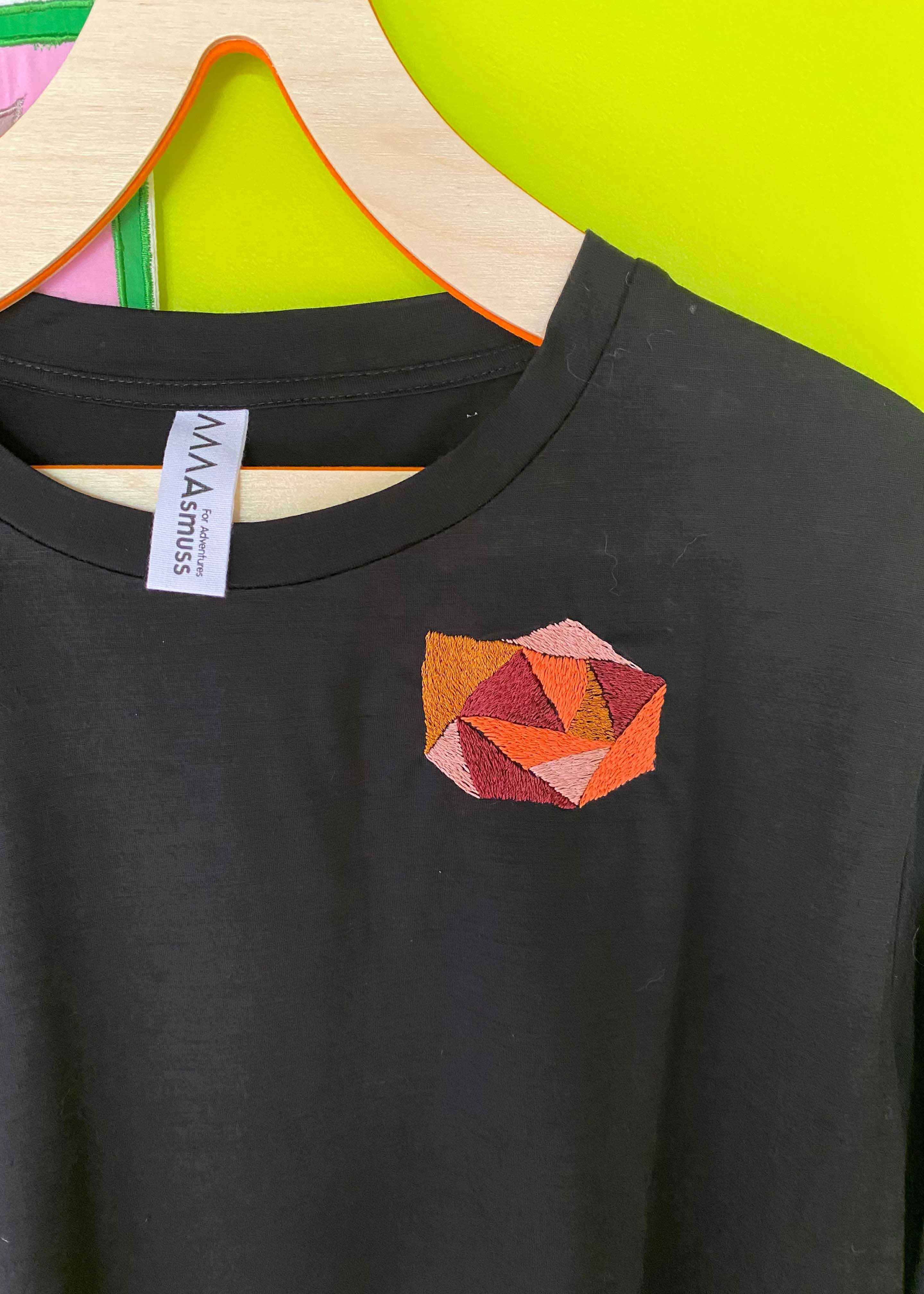 close up of hand embroidery on black tee shirt. Hand embroidery is an abstract geometric rose in oranges, mustard, deep red and pale pink